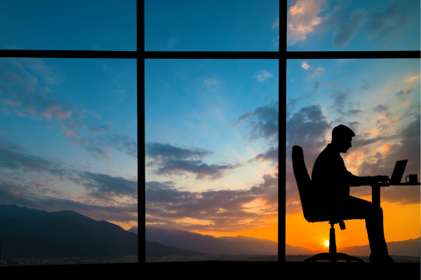 view of sunset through large windows, with silhouette of person on laptop in bottom corner 