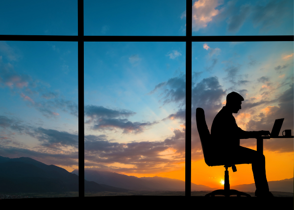 view of sunset through large windows, with silhouette of person on laptop in bottom corner 