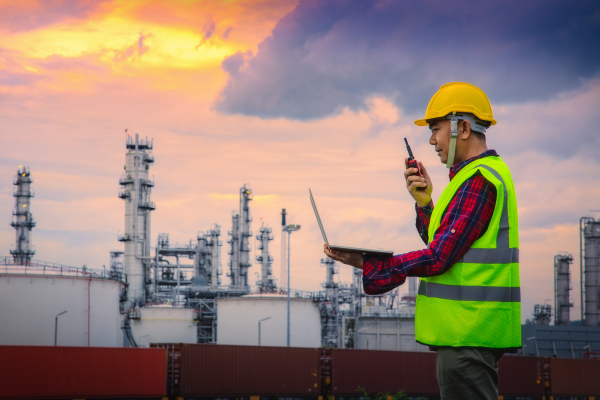 person in hard hat and reflective vest standing in front of oil rig holding a laptop