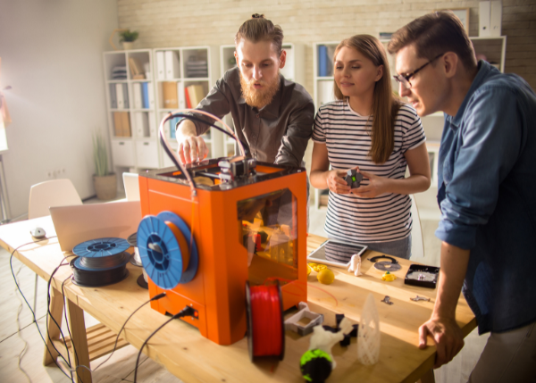 three people watching 3D printer with tools on table around them