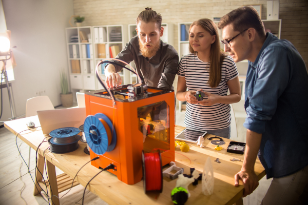 three people watching 3D printer with tools on table around them