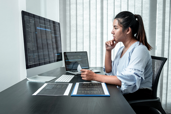 woman sitting at table with multiple computer screens and papers 