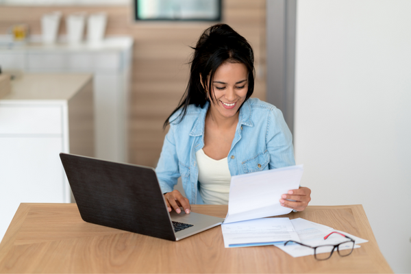 woman seated at table with laptop and holding piece of paper
