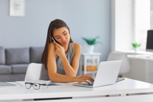 Female at home desk, typing on laptop and holding phone to ear