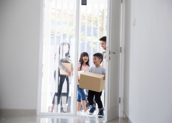 Parents and two children smiling, carrying moving boxes, entering vacant apartment