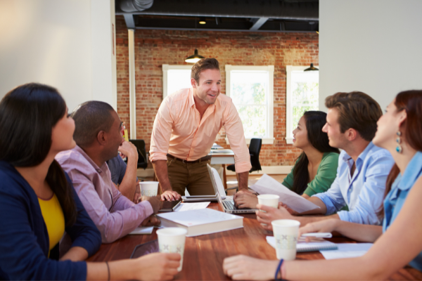 Casual man standing front of table leaning towards sitting employees